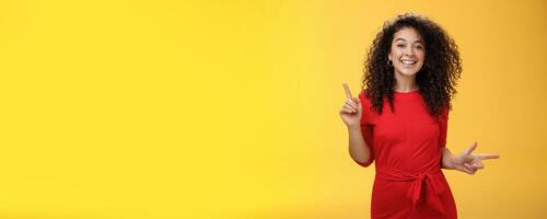 Charismatic cute and happy young curly-haired european female in red dress dancing and pointing up and right, showing sideways as presenting choices picking, making decision and smiling at camera photo