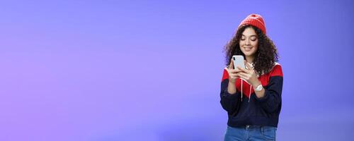 Woman searching in internet funny memes, holding mobile phone in hands smiling joyfully and cute at smartphone screen, typing message or browsing internet over blue background, wearing warm winter hat photo