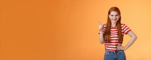 Cheerful sly redhead girlfriend have something interesting on mind, squinting cunning joyful smile, hold hand waist confident pose, pointing upper left corner give recommendation, orange background photo