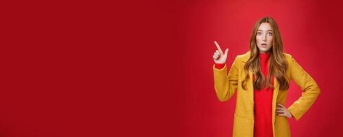 Shocked and stunned cute, confused ginger girl in yellow coat feeling worried and surprised open mouth lifting eyebrows amazed pointing at upper left corner, posing against red background photo