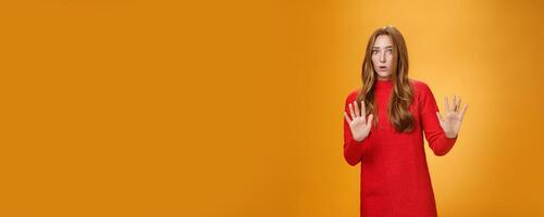 Intense panicking and freaked out girl with red hair asking slow down, raising hands near chest in stop and not gesture open mouth surpised and shocked, dismaying strange offer over orange background photo