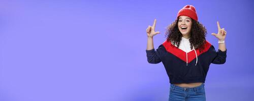 Waist-up shot of cheerful energized young woman calling friend inviting hang out as weather awesome pointing up with raised hands smiling broadly posing in red winter hat over blue background photo