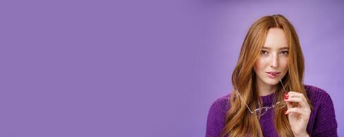 Close-up shot of smart and creative female redhead genius biting frame of glasses and squinting curious at camera as thinking having idea, making up plan against purple background photo
