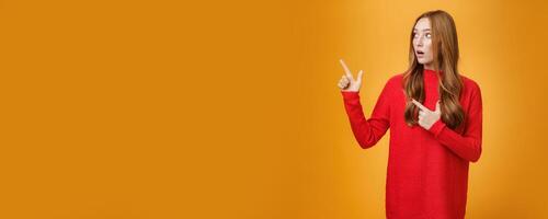 Impressed and curious good-looking stylish 20s redhead woman with cute curls in red dress drop jaw from amazement and interest as looking and pointing at upper left corner speechless photo