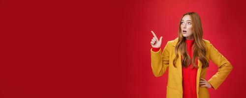 Portrait of empathic shocked and surprised stunned redhead woman in yellow coat open mouth amazed and speechless pointing and looking at upper left corner concerned and shook over red wall photo