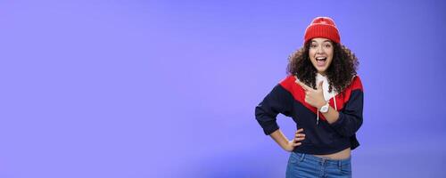 Portrait of enthusiastic and sociable european woman in warm hat and sweatshirt smiling delighted with amused grin as pointing at upper left corner impressed and astonished with awesome promotion photo