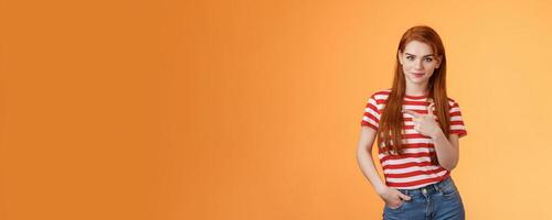 Click right now. Confident sassy good-looking redhead woman smiling cunning self-assured, pointing left, showing copy space promo, grinning empowered, know what best, orange background photo
