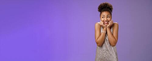 Amused silly cute black girl in silver dress anticipating exciting party dreamy gaze up touch cheeks smiling broadly optimistic waiting dream come true standing pleased touched blue background photo