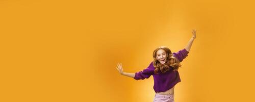 Expressive and enthusiastic carefree friendly joyful redhead woman in purple sweater and pants jumping joyfully with raised hands smiling yelling from happiness and joy against orange background photo