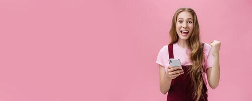 Yes I did it. Happy young excited cute woman in trendy outfit expressing cheer and joy smiling happily at camera raising fist in triumph and success holding smartphone beating own record in game photo