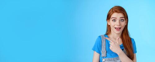 Curious excited redhead woman attend interesting exhibition, pointing left look camera interested, questioned about copy space promo, asking question intrigued cool offer, blue background photo