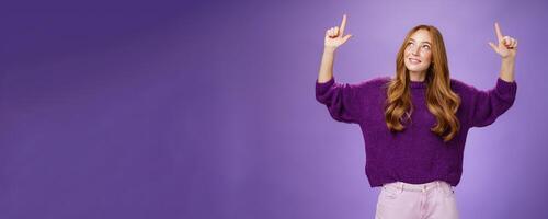 Dreamy girl with red hair and freckles in warm cozy purple sweater raising hands looking and pointing up with intrigued and happy charmed expression smiling as liking curious product over violet wall photo