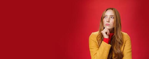 Close-up shot of determined and focused creative thoughtful redhead female looking at upper left corner touching chin, thinking, making choice or remembering information over red background photo