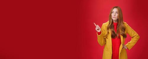 Sad and gloomy cute ginger girl in 20s wearing trendy yellow coat holding hand on waist, pouting, looking and pointing at upper left corner with regret and sorrow over red background photo