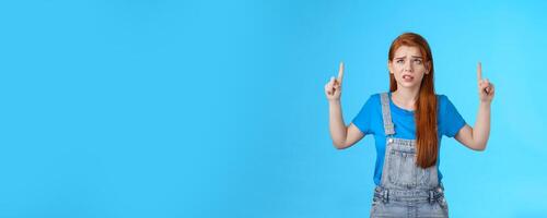 Uneasy worried cute redhead female coworker feel unsure insecure, grimacing displeased intense worried expression, look pointing up, top copy space, have hesitations, stand blue background photo