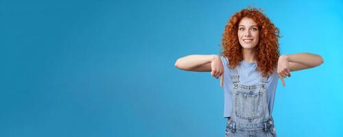 Excited happy smiling caring redhead curly-haired girlfriend thrilled awaiting summer holiday trip pointing down index finger upbeat mood show favorite store buy best prices, blue background photo