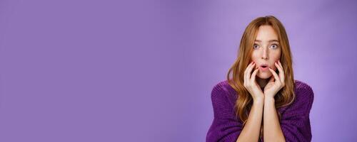 Shocked redhead female friend touching face and open mouth as looking with widen eyes at camera, being overwhelmed with shook and amazement hearing terrible story over purple background photo
