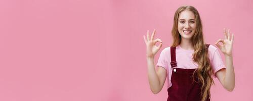 No problem, everything okay. Portrait of pleasant and kind pretty young female in dungarees showing ok or perfect gesture with both hands smiling broadly with friendly gaze at camera over pink wall photo