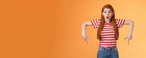 Shocked excited attractive redhead woman gasping, drop jaw shook, stare camera astonished, pointing down, feel amazed and surprised see incredible good offer, gossiping stunned, orange background photo