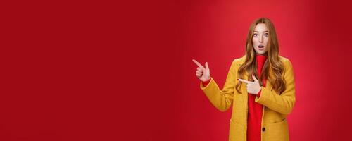 Stunned speechless and impressed attractive redhead woman with freckles in yellow coat drop jaw amazed as popping eyes at camera questioned, pointing left astonished over red background photo