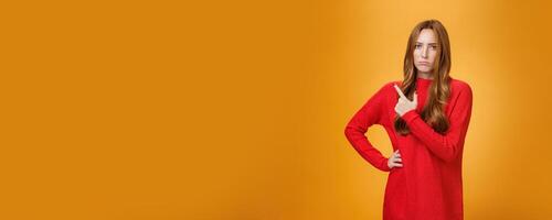 Upset and disappointed young ginger girl in red sweater pointing at upper left corner or behind frowning looking gloomy and angry, standing jealous and moody against orange background photo
