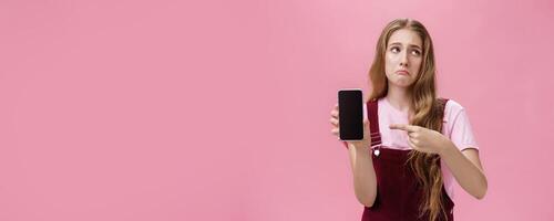 Indoor shot of gloomy displeased and disappointed cute young girl holding smartphone pointing at cellphone screen making upset grimace as if being bothered and dissatisfied against pink wall photo