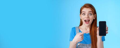 Excited amused good-looking ginger girl promote awesome app, show smartphone display, pointing telephone screen astonished, smiling surprised, like cool game, stand blue background photo