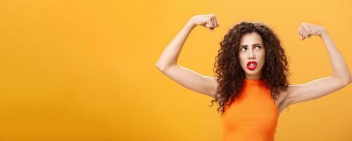 Woman feeling powerful and strong raising hands with clenched fists making intense face being working out in gym showing muscles and biceps looking at upper right corner posing over orange background photo