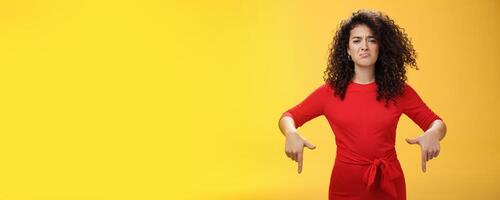 Gloomy disappointed cute silly curly-haired female in red dress frowning upset and making sad smile as pointing down with regret and unsatisfied expression, jealous or displeased over yellow wall photo