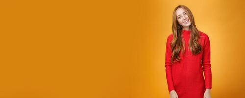 Attrative and stylish elegant busiensswoman with ginger hair in red knitted dress tilting head and smiling flirty and happily posing over orange background as listening with interest to conversation photo