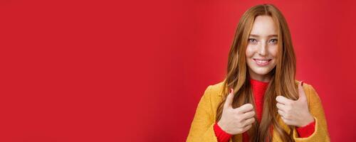 Close-up shot of satisfied happy young 20s woman in yellow coat showing thumbs up gesture in like and approval giving positive feedback enjoying nice event standing over red background, smiling cute photo