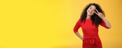 Waist-up shot of friendly tender and feminine cute woman with curly hairstyle tilting head and smiling, laughing and showing victory or peace gesture at camera over yellow background photo