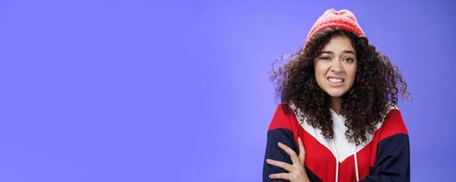 Woman with curly hair in winter beanie feeling uncomfortable and discomfort clenching teeth and frowning intense as hugging herself insecure and awkward, unwilling to say cruel rejection, feel awkward photo