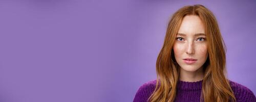 Headshot of sensual and attractive tender redhead female with freckles and pure skin looking at camera focused and sincere with slightly opened mouth, posing against purple background photo