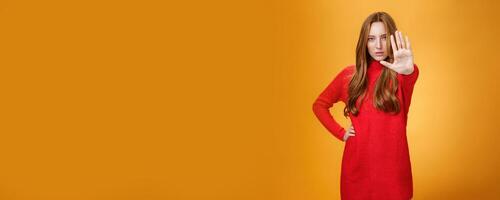 Confident sensual and attractive redhead female with self-assured gaze extending palm towards camera in stop and prohibition gesture demanding halt, stand still over orange background photo