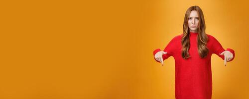 Displeased serious-looking female in knitted red dress frowning looking concerned as pointing down at displeasing unappropriate thing standing disappointed and unhappy over orange background photo