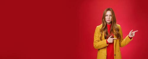 Angry and offended disappointed redhead female in yellow coat frowning looking strict and serious at camera with dislike and agression pointing right asking explain yourself over red background photo