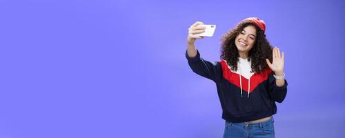 Studio shto of stylish cute female vlogger with curly hair in winter red hat smiling and waving hello at smartphone camera as recording , making interesting content to post online over blue wall photo