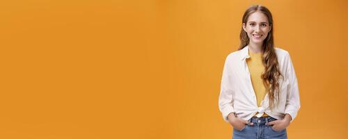 Stylish young female designer wants help friend shopping holding hand in pockets smiling joyfully and self-assured at camera wearing trendy blouse over yellow t-shirt posing over orange wall photo