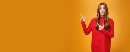 Surprised and impressed intrigued nice cute redhead female with freckles in warm red sweater pointing at upper left corner open mouth from amazement and looking questioned at camera over orange wall photo