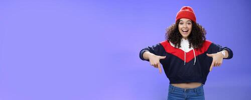 Hurry up and chek it out. Portrait of charismatic joyful and excited young cute curly-haired girl in beanie and stylish outfit pointing down, smiling broadly inviting join fun over blue background photo