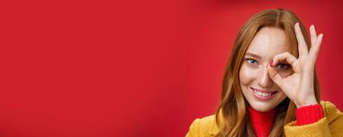 Zero worries only happiness. Portrait of charming cute and carefree redhead female with freckles and blue eyes smiling delighted showing okay gesture on eye and peeking at camera over red background photo