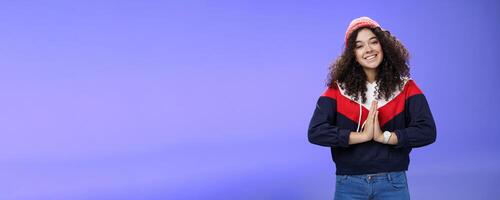 Portrait of charming curly-haired feminine girlfriend begging for favour as posing over blue background in winter outfit with hat, holding hands in pray smiling with angel look and friendly gaze photo