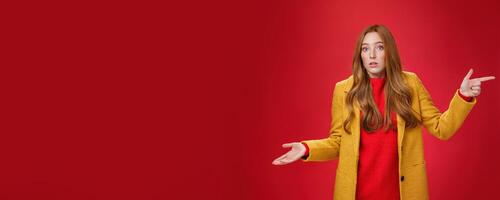 Woman missed client posing confused and unaware how happened shrugging making clueless gesture with palm as pointing right and looking questioned and uncertain at camera over red background photo