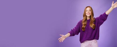 Girl stretched hands sideways to welcome and greet friend giving warm hug smiling broadly at camera standing joyful wanting cuddle over purple background, wearing violet sweater and pants photo