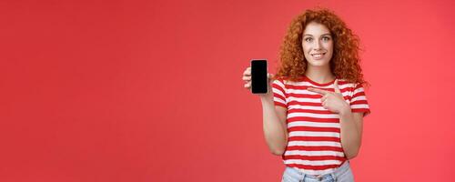 How about this app. Redhead curly friendly cute girl help friend find oufit online store hold smartphone pointing phone screen smiling broadly recommend social media photo filter, red background
