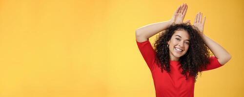 Close-up shot of charismatic playful and tender young kind woman with curly hair playing having fun showing bunny ears with hands on head tilting head joyfully fooling around and smiling at camera photo