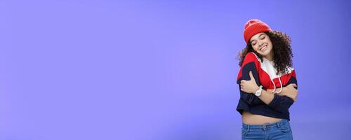 Sweet tender and sensual curly-haired woman in red stylish beanie hugging herself with closed eyes and nostalgic smile leaning on shoulder feeling warmth and tenderness as recalling loving memories photo