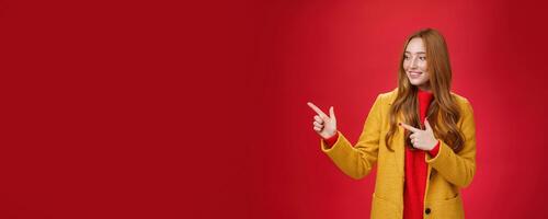 Portrait of curious and happy pleased attractive redhead woman in yellow coat pointing and looking left with delighted intrigued smile observing awesome copy space against red background photo
