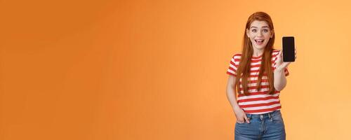 Excited cute outgoing redhead female impressed showing app, hold smartphone introduce gadget feature, smiling surprised, look enthusiastic like application, brag game score, orange background photo